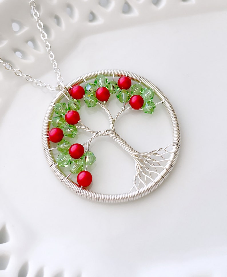 Op de afbeelding: Een zilveren draadboom des levens ketting met rode en groene kralen. De ketting ligt op een witte achtergrond.