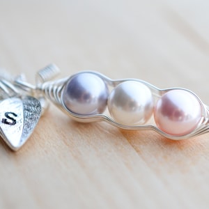 May include: A silver necklace with a charm shaped like a pea pod with three pearls inside. The pearls are lavender, white, and pink. The necklace also has a small silver heart charm with the letter 'S' engraved on it.