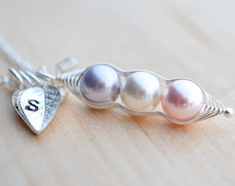 May include: A silver necklace with a charm shaped like a pea pod with three pearls inside. The pearls are lavender, white, and pink. The necklace also has a small silver heart charm with the letter 'S' engraved on it.