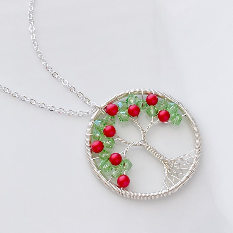 Op de afbeelding: Een zilveren draadgewikkelde levensboomketting met rode en groene glazen kralen. De ketting ligt op een witte achtergrond.