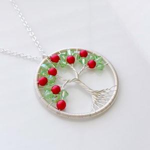 Op de afbeelding: Een zilveren draad gewikkelde boom des levens ketting met rode en groene kralen. De ketting ligt op een witte achtergrond.
