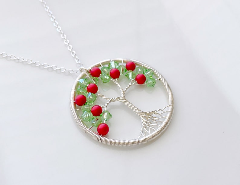 Op de afbeelding: Een zilveren draad gewikkelde boom des levens ketting met rode en groene kralen. De ketting ligt op een witte achtergrond.