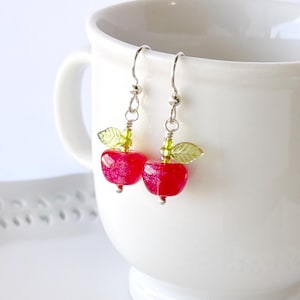 May include: A pair of apple-shaped earrings. Each earring features a faceted red bead, a small green leaf, and a silver-colored hook. The earrings are hanging in front of a white mug.