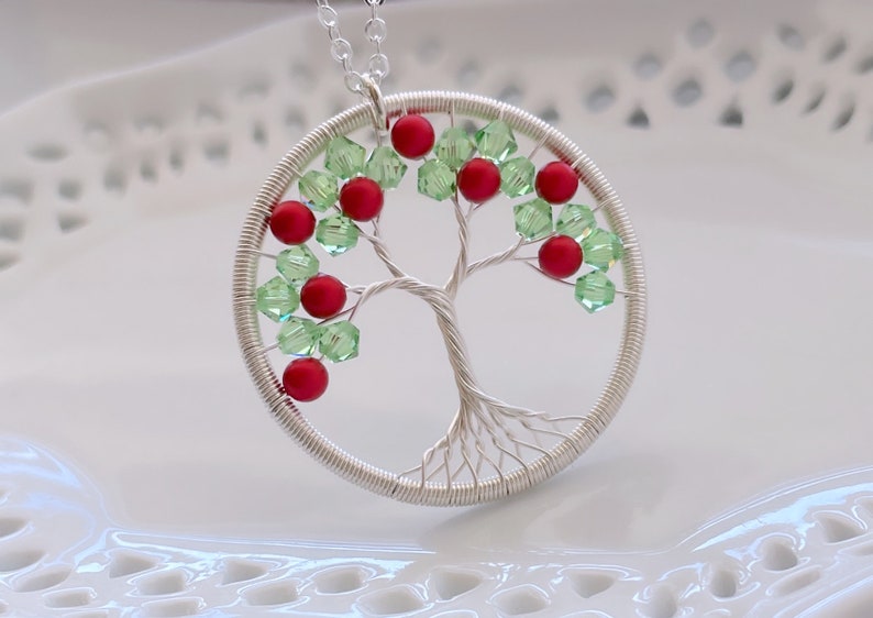 Op de afbeelding: Een zilveren draadgewikkelde boom des levens hanger ketting met rode en groene kralen. De hanger is rond en hangt aan een zilveren ketting.