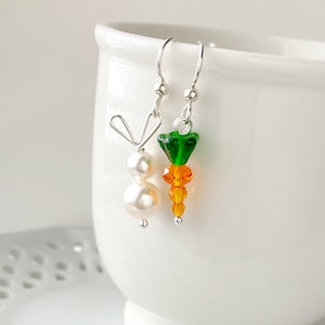 May include: A pair of dangle earrings featuring a carrot and a bunny. The carrot is made of orange and green glass beads. The bunny is made of white pearl beads and silver wire.