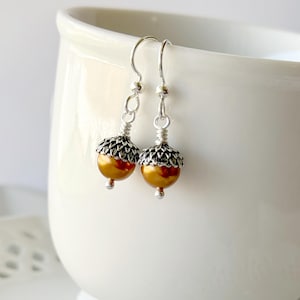 May include: A pair of acorn-shaped earrings. The earrings feature silver-colored hooks and caps, with a detailed, textured design. The acorn bodies are a warm, golden-brown color with a hint of red. The earrings are displayed against a white background.