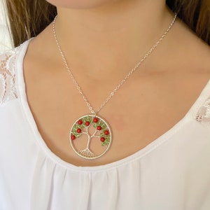 Op de afbeelding: Een zilveren ketting met een levensboom hanger. De hanger is gemaakt van draad en heeft groene en rode kralen. De ketting ligt op een wit shirt.