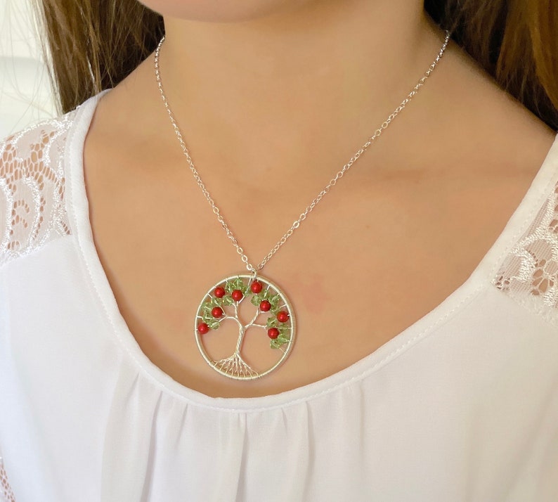 Op de afbeelding: Een zilveren ketting met een levensboom hanger. De hanger is gemaakt van draad en heeft groene en rode kralen. De ketting ligt op een wit shirt.