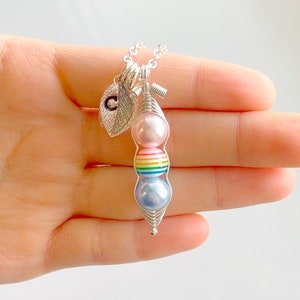 May include: A silver necklace with a charm of a pea pod with three beads. The beads are pink, rainbow striped, and light blue. The necklace also has a silver charm shaped like a leaf with the letter C on it.