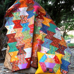 May include: A colorful patchwork quilt with a geometric pattern of stars and triangles. The quilt is made of various fabrics with different colors and patterns. A smaller pillow with a similar geometric pattern is in front of the quilt.