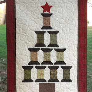 May include: A white quilted wall hanging with a red border features a Christmas tree design made from brown spools of thread. The spools are arranged in a pyramid shape with a red star on top.