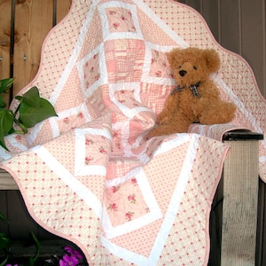以下が含まれることがあります： ピンクと白のパッチワークのキルトで、フリルのある縁取りが施されています。花柄のプリントが施され、その上にテディベアが座っています。