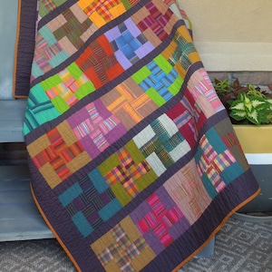 Może przedstawiać: Kolorowa kołdra w patchworkowym wzorze, z różnymi tkaninami w kratę w odcieniach zieleni, błękitu, czerwieni, pomarańczy i fioletu. Kołdra składa się z kwadratów z wzorem krzyża w każdym kwadracie. Kołdra jest złożona i położona na ławce.