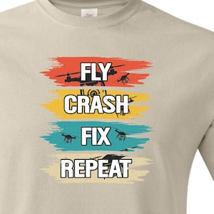 May include: A beige t-shirt featuring the text "FLY, CRASH, FIX, REPEAT" in a stacked format. Each word is on a different coloured horizontal stripe: red, yellow, teal, and beige. The shirt also has the words "Natural / Canvas" at the bottom.