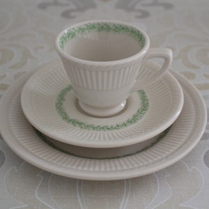 May include: A white teacup and saucer set with a green floral design. The saucer has a raised edge and a scalloped design.
