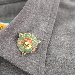 May include: A circular metal lapel pin featuring a green border with the text "SKI APACHE" and "ELEV. 11,500". The center displays a red sunburst over a mountain silhouette and two pinecone icons, all set against a triangular gold-toned backing.