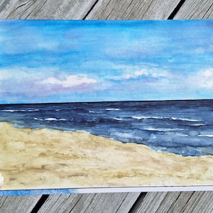 Könnte beinhalten: Aquarellmalerei einer Strandszene mit blauem Himmel, Ozean und Sandstrand. Das Kunstwerk zeigt Blautöne, Beige und Weiß. Muscheln sind um das Gemälde angeordnet. Ein Pinsel mit grünem Griff liegt auf der Holzoberfläche.