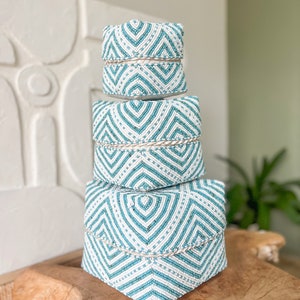 Könnte beinhalten: Drei dekorative Boxen, die übereinander gestapelt sind. Sie sind mit weißen und türkisfarbenen Perlen in einem geometrischen Muster bedeckt. Jede Box hat ein helles, gewebtes Band. Die Boxen stehen auf einer Holzoberfläche.