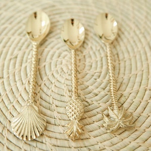 May include: Three gold-tone spoons with different decorative handles. The handles are shaped like a seashell, a pineapple, and a palm tree.