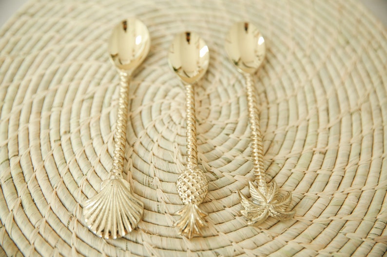 May include: Three gold-tone spoons with different decorative handles. The handles are shaped like a seashell, a pineapple, and a palm tree.