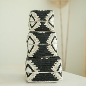 Könnte beinhalten: Ein Stapel von drei quadratischen Aufbewahrungsboxen. Die Boxen sind schwarz mit geometrischen Mustern aus weißen Perlen. Die Boxen sind unterschiedlich groß, die kleinste oben und die größte unten.