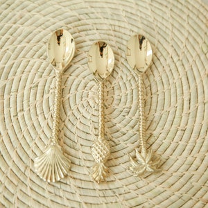 May include: Three gold-toned spoons with decorative handles. The handles are shaped like a seashell, a pineapple, and a palm tree. The spoons are on a woven straw placemat.
