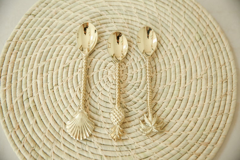 May include: Three gold-toned spoons with decorative handles. The handles are shaped like a seashell, a pineapple, and a palm tree. The spoons are on a woven straw placemat.