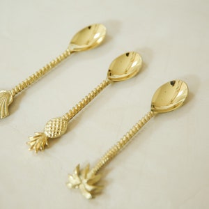 May include: Three gold-tone spoons with decorative handles. The handles are shaped like a seashell, a pineapple, and a palm tree.