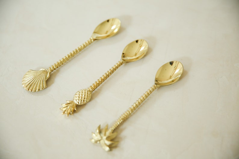 May include: Three gold-tone spoons with decorative handles. The handles are shaped like a seashell, a pineapple, and a palm tree.