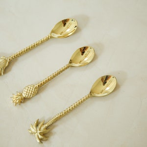 May include: Three gold-toned spoons with different tropical-themed handles. The first spoon has a seashell handle, the second has a pineapple handle, and the third has a palm tree handle.