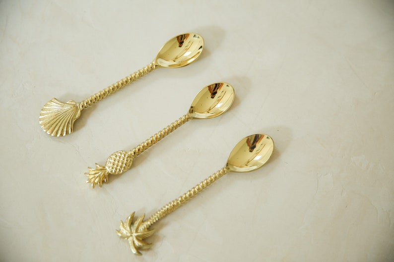 May include: Three gold-toned spoons with different tropical-themed handles. The first spoon has a seashell handle, the second has a pineapple handle, and the third has a palm tree handle.