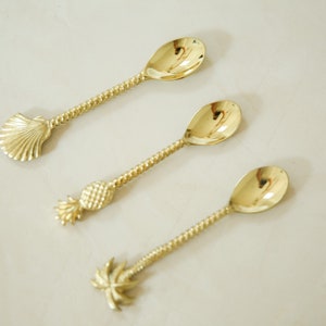 May include: Three gold-tone spoons with different tropical-themed handles. The first spoon has a seashell handle, the second has a pineapple handle, and the third has a palm tree handle.