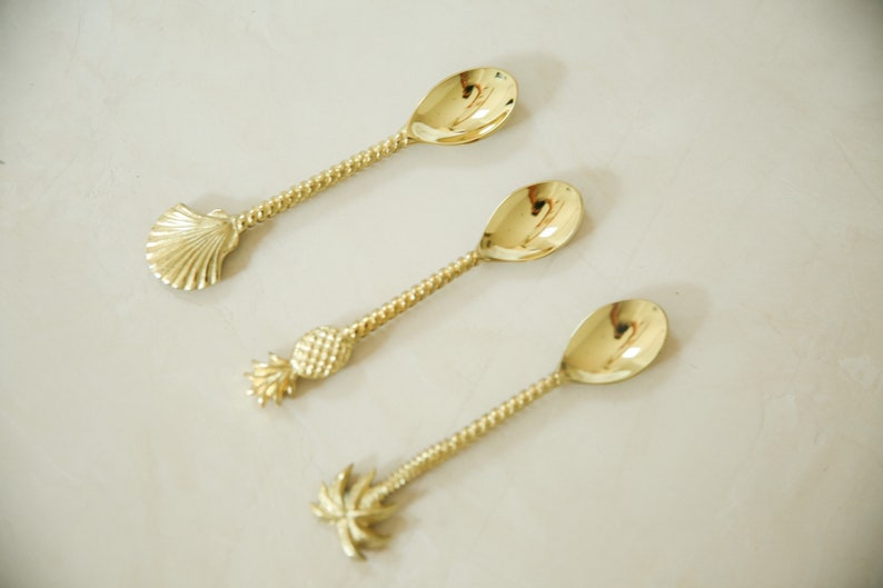 May include: Three gold-tone spoons with different tropical-themed handles. The first spoon has a seashell handle, the second has a pineapple handle, and the third has a palm tree handle.