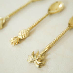 May include: Three gold-tone spoons with different tropical-themed handles: a seashell, a pineapple, and a palm tree.