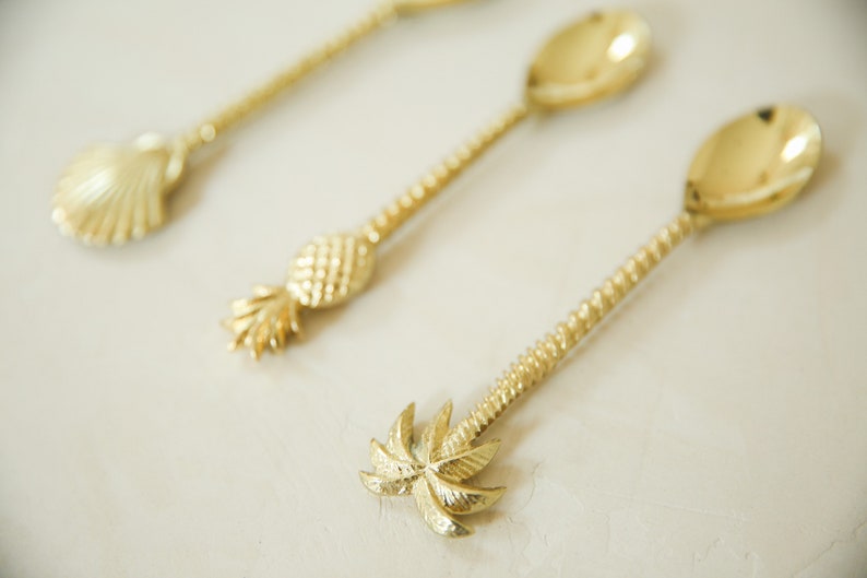 May include: Three gold-tone spoons with different tropical-themed handles: a seashell, a pineapple, and a palm tree.