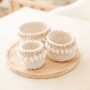 Op de afbeelding: Drie witte gehaakte manden met een schelpbezetting op een houten dienblad. De manden zijn van verschillende grootte en zijn in een rij gerangschikt.