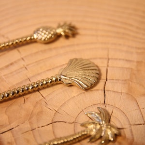 May include: Three gold-toned stirrers with different tropical designs: a pineapple, a seashell, and a palm tree. The stirrers are on a wooden surface.