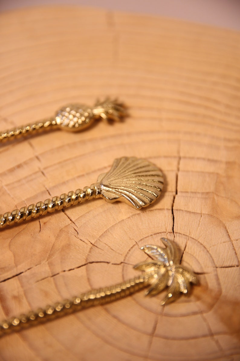 May include: Three gold-toned stirrers with different tropical designs: a pineapple, a seashell, and a palm tree. The stirrers are on a wooden surface.