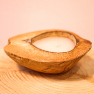 Puede incluir: Un portavelas de madera con forma de media cáscara de coco con una vela blanca en su interior. El portavelas está sobre una superficie de madera.