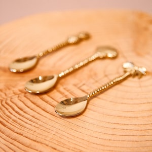 Puede incluir: Tres cucharas doradas con un diseño retorcido. Las cucharas están sobre una superficie de madera.