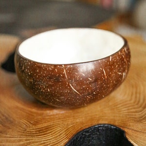 Puede incluir: Cuenco de coco hecho a mano con exterior marrón natural e interior blanco. El cuenco está sobre una superficie de madera, mostrando la veta y textura natural de la madera. Este cuenco ecológico es ideal para servir alimentos o como pieza decorativa.