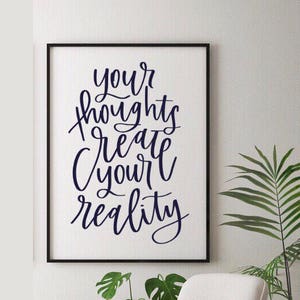May include: A black framed print with a white background and the text "Your thoughts create your reality" in blue cursive lettering.