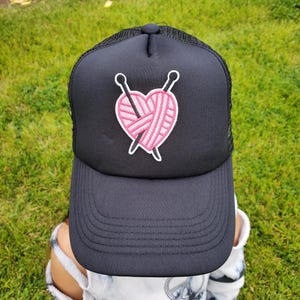 May include: A black trucker hat with a pink heart and knitting needles embroidered on the front.