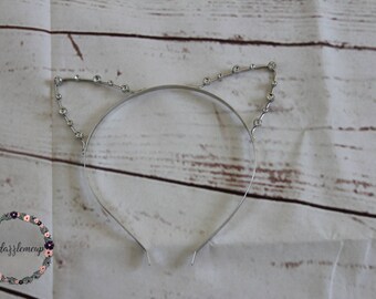 May include: A silver headband with rhinestones forming cat ears. The headband is on a white wooden background.