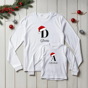 May include: Two white long-sleeved shirts, one adult and one child size, with Christmas designs. The adult shirt has a black "D" with "Daddy" and a Santa hat. The child's shirt features a black "A" with "Anabelle" and a Santa hat, festive for the holidays.