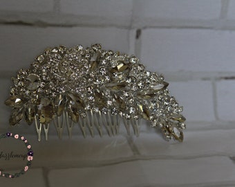 Puede incluir: Un peine de pelo plateado con un gran grupo de diamantes de imitación transparentes y dorados. El peine está decorado con un diseño floral y tiene una fila de dientes en la parte inferior.