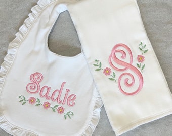 May include: White baby bib and burp cloth set. The bib features the name "Sadie" embroidered in pink with floral accents. The burp cloth has a pink embroidered "S" with floral details. Both items have a white background.