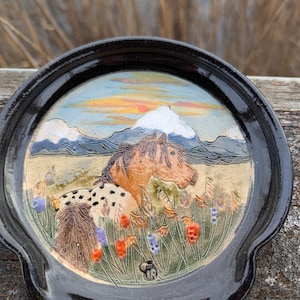 May include: A black ceramic spoon rest with a hand-painted horse scene. The design features a brown and white horse in a field with colorful flowers, mountains, and a sunset. The spoon rest has a curved handle.