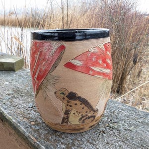 May include: A ceramic vase with a brown and black glaze. The vase is decorated with a hand-painted design of a frog sitting under a red mushroom.