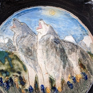 May include: A ceramic plate with a hand-painted design of two wolves howling at the moon. The wolves are painted in shades of gray and white, and the background is a blue sky with white clouds. The plate is decorated with a green and brown border.